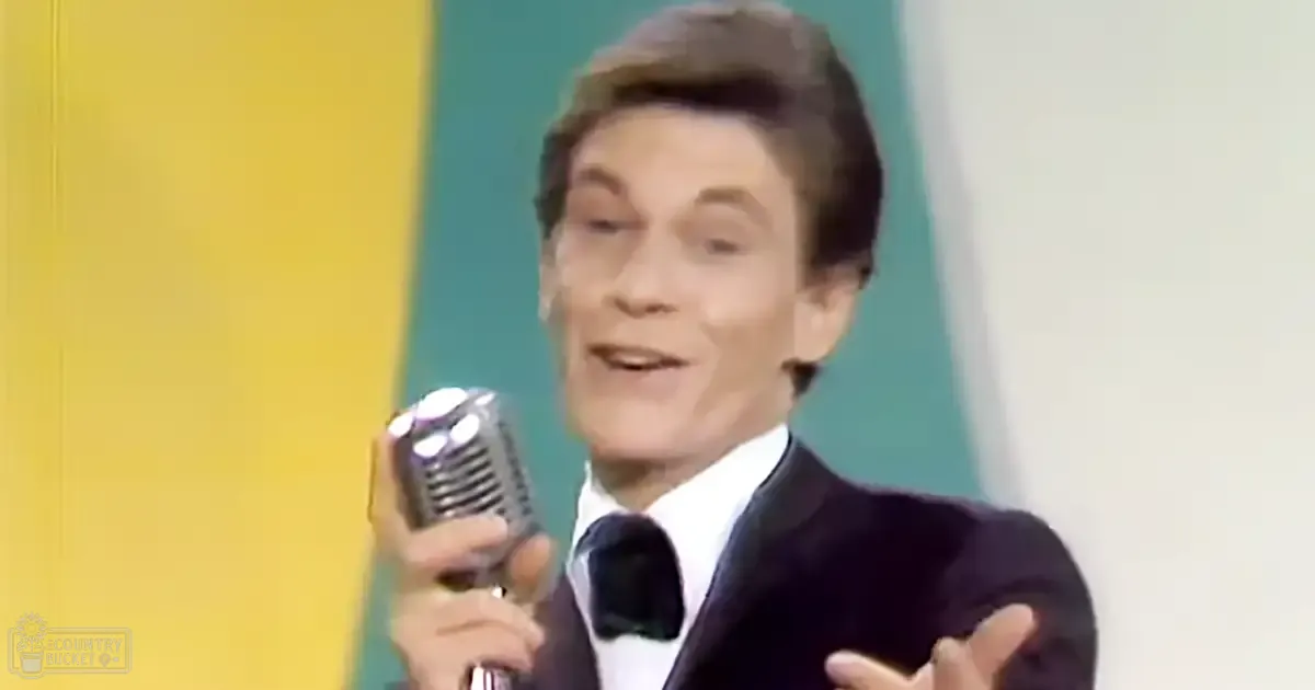 An adult male performer in a mid-20th century suit, holding a vintage microphone on stage with yellow and teal panels behind him.