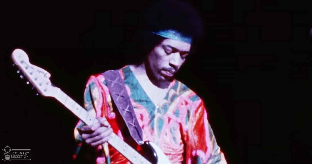 A male adult musician in a colorful, patterned 1960s-style garment plays an electric white guitar on stage under focused lighting.
