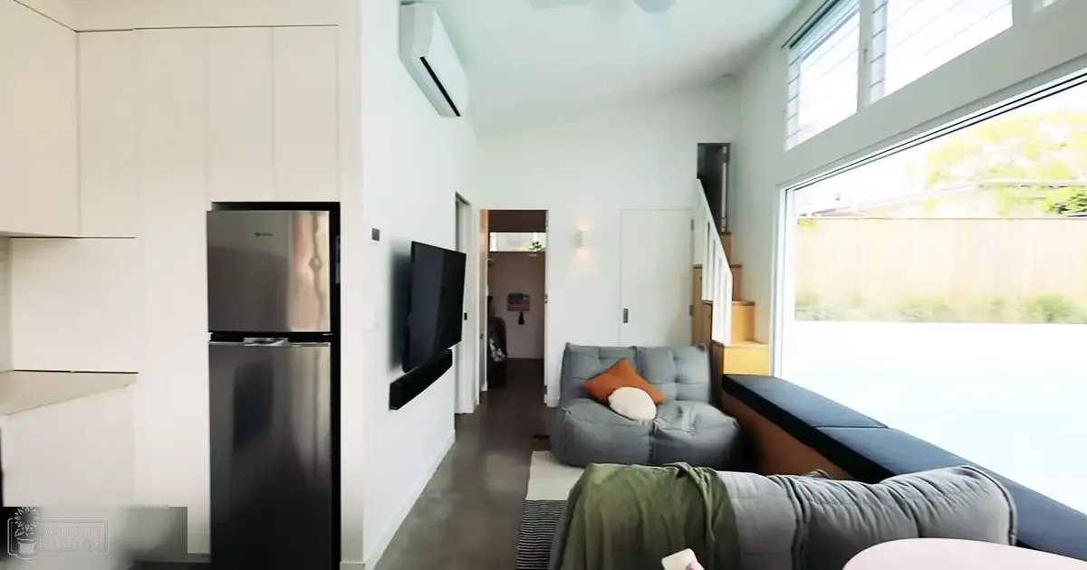 Modern minimalist living space with large windows, blue sofa, orange cushions, green blanket, stainless steel refrigerator, wooden staircase, spring or summer foliage outside