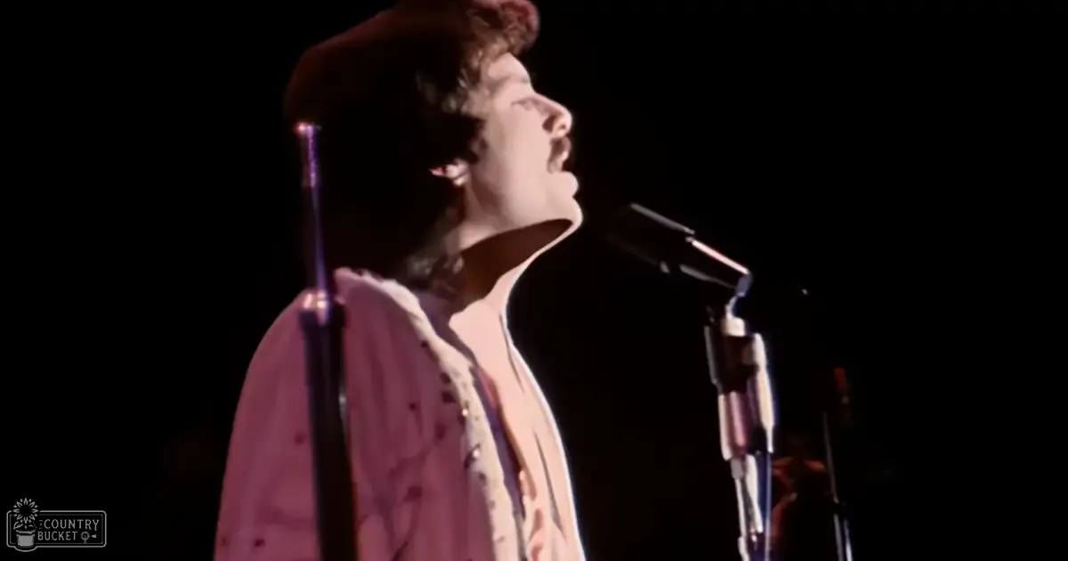 Young adult male in a relaxed, casual style open-collared shirt with an abstract pattern on stage at an indoor concert venue under spotlights.