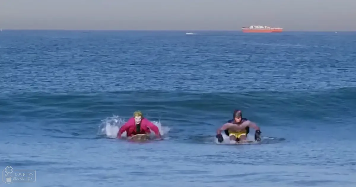Two young adult males surfing in ocean waves at a beach, with calm skies and a distant ship visible.