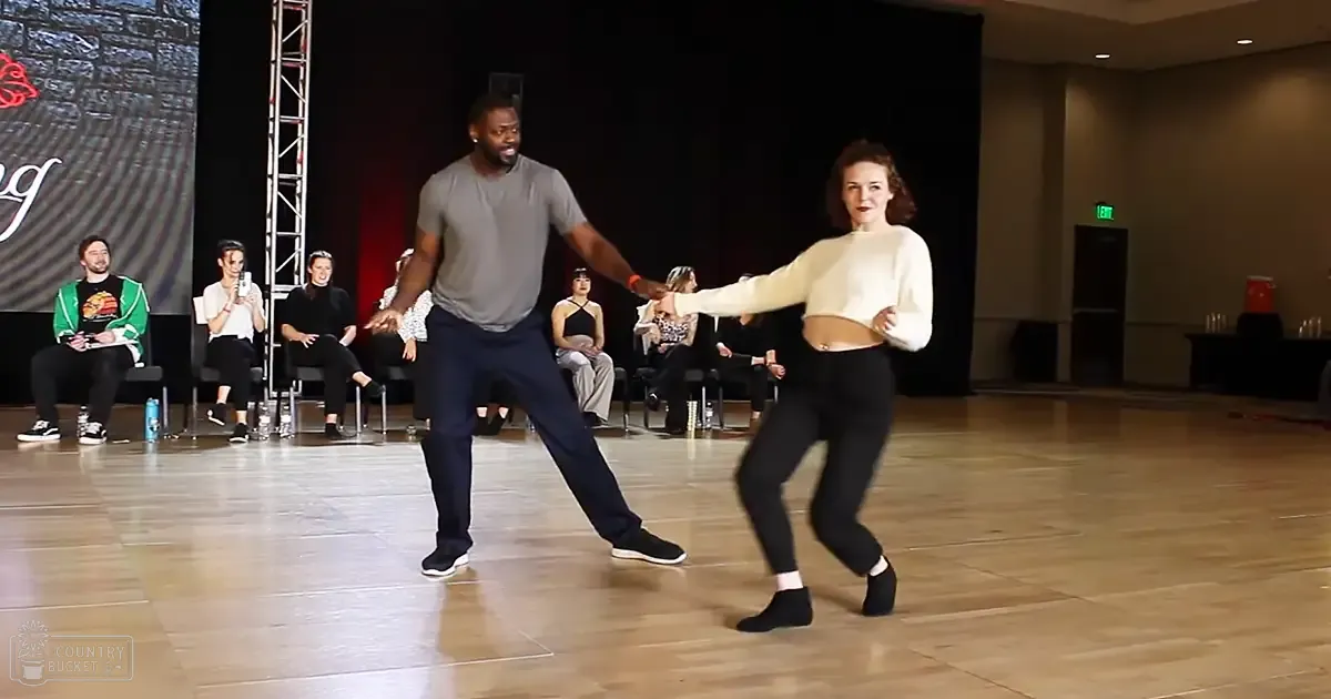 An adult male and female dancing in a dance studio with spectators seated around. The setting is indoors, likely during evening hours.