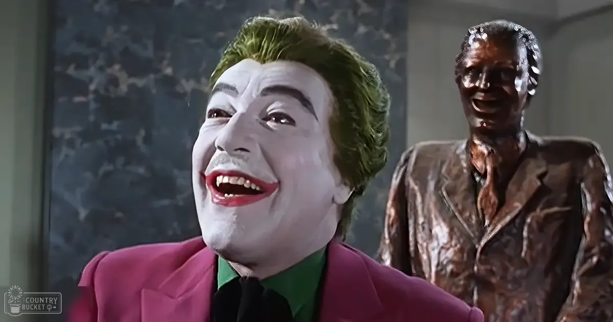 An adult male with green hair styled upwards, wearing a bright pink suit jacket over a dark shirt and a green collar, set against a backdrop of a patterned wall in an indoor theatrical or film setting.