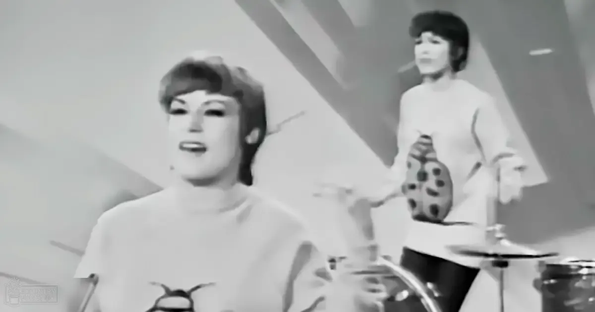 An adult female singer wearing a light-colored top with a rounded neckline and short sleeves, accompanied by another adult female drummer in a white dress or tunic with bell-shaped sleeves, set against an indoor stage backdrop.