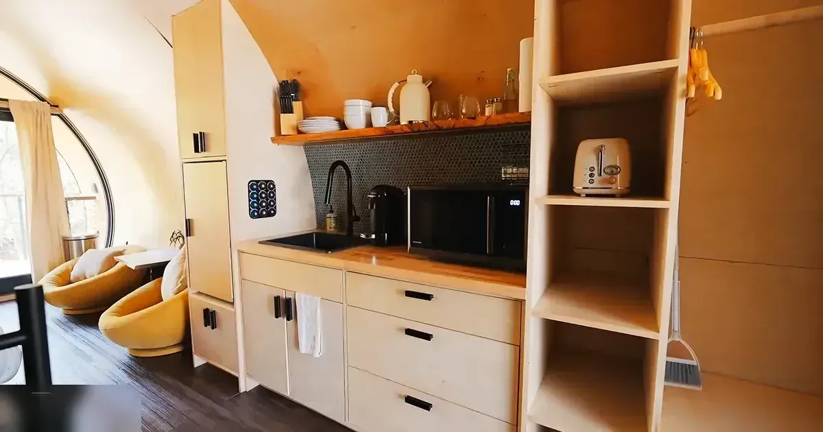 Compact kitchenette with light-colored cabinets, microwave, sink, utensils, dishes, kettle, toaster, broom, and two yellow armchairs in a modern, possibly tiny home or cabin setting.