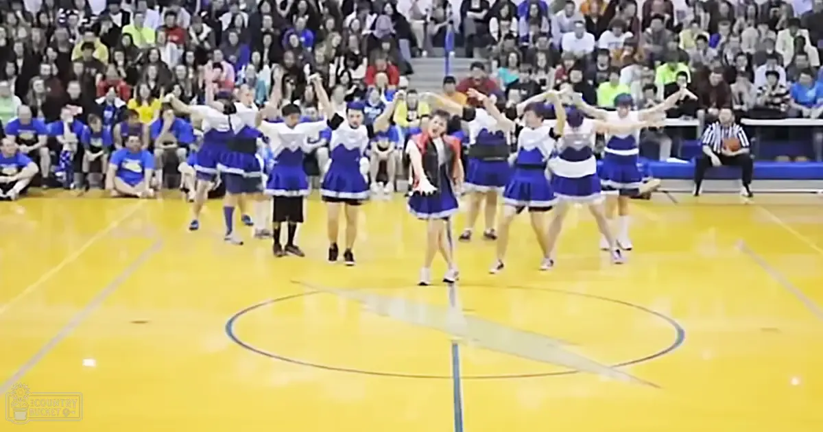 A cheerleading team in blue and white uniforms performs at an indoor sports arena filled with spectators during a daytime event.