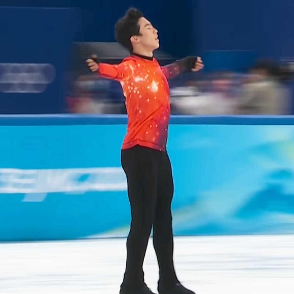 A young adult male figure skater in a red, snowflake-patterned skating outfit at an indoor ice rink with blue walls.