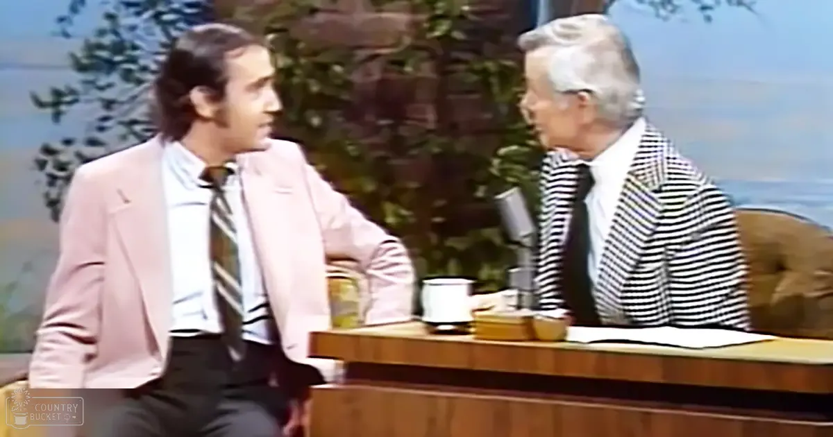 Two adult males in a studio setting, one wearing a light pink blazer and striped tie, the other in a black and white striped blazer over a white shirt. A wooden desk with mugs and papers is visible on stage.