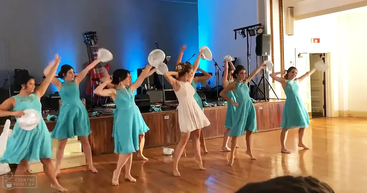 Young adult female dancers in turquoise and white sleeveless dresses perform on a wooden stage with blue ambient lighting, surrounded by musical instruments under indoor venue conditions.