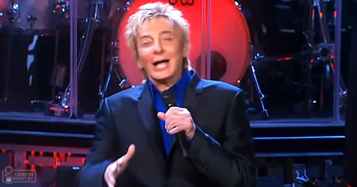 An adult male performer in a dark suit jacket and blue shirt on an indoor stage with musical instruments, wearing 'Madly Odd' text on clothing.
