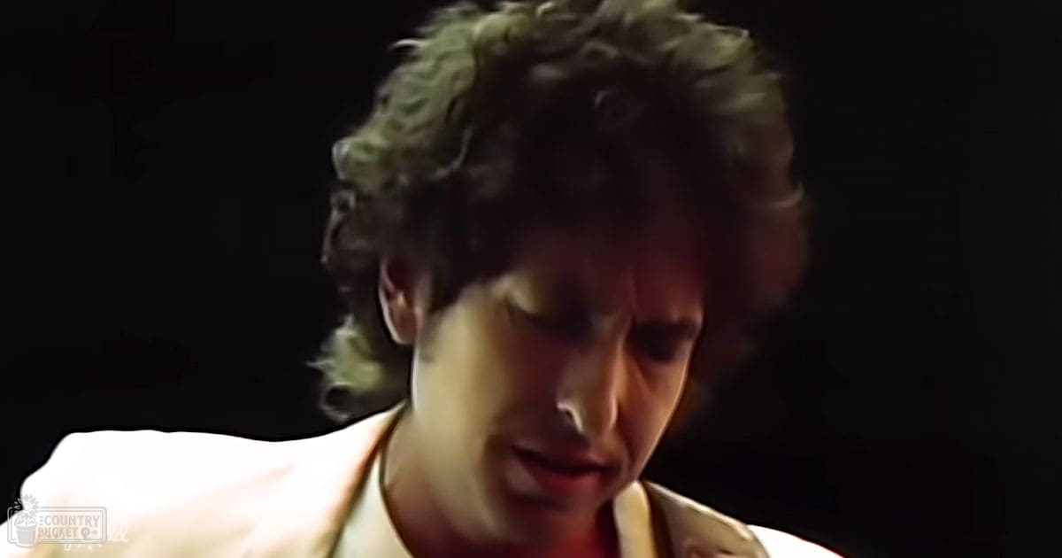 A young adult male in a dark indoor setting holding a light-colored garment.