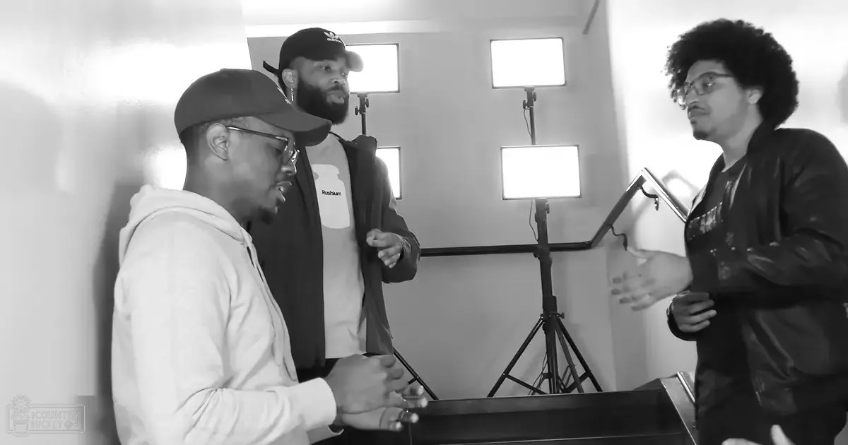 A group of adult males in a studio setting with modern casual attire, including hoodies, jackets, and a microphone stand visible behind them.