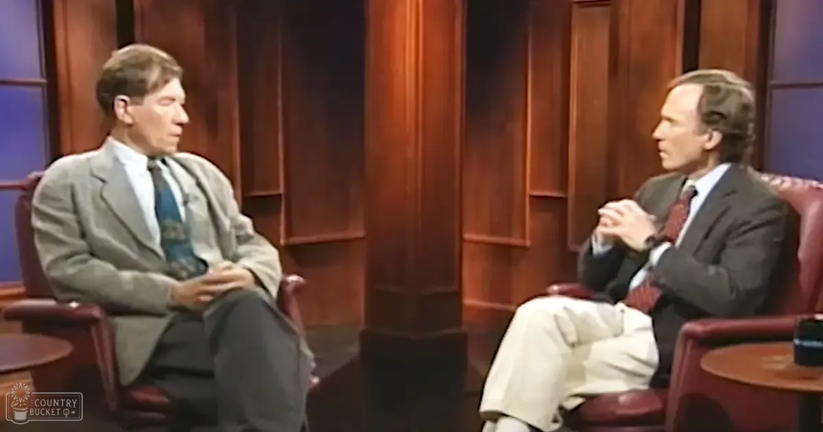 Two adult males engaged in conversation in a formal indoor studio setting with maroon leather armchairs, wooden paneling, and a dark blue accent wall.