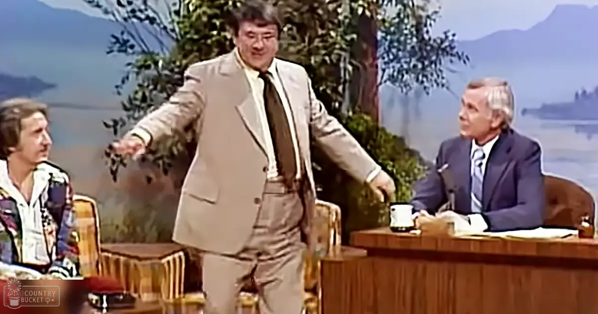 A man mid-gesture on a talk show set, surrounded by other adults in suits, against an outdoor scenic backdrop.