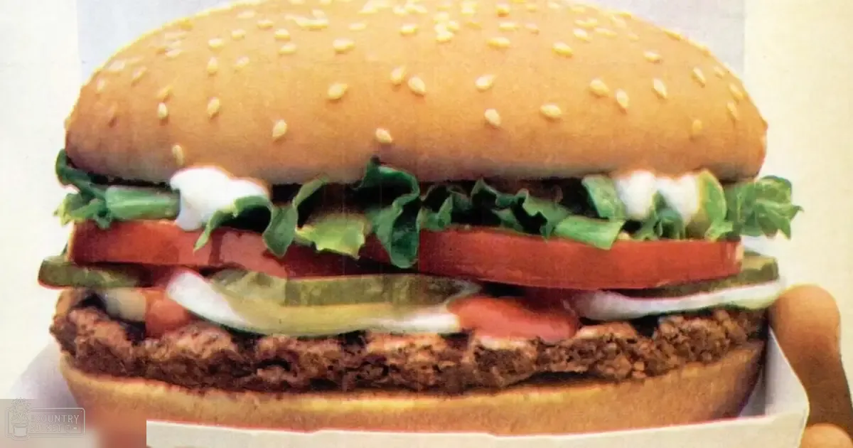 Close-up of a hamburger with sesame seed buns, lettuce, tomato slices, pickles, onions, and creamy sauce in an indoor setting.