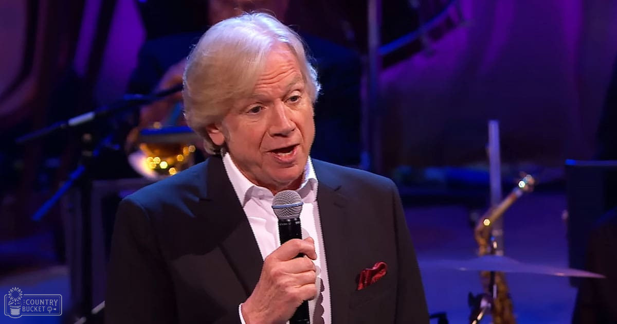 An older adult male singer performing on a dimly lit indoor stage with purple lighting, wearing a dark suit jacket and white shirt with a red pocket square, surrounded by musical instruments.