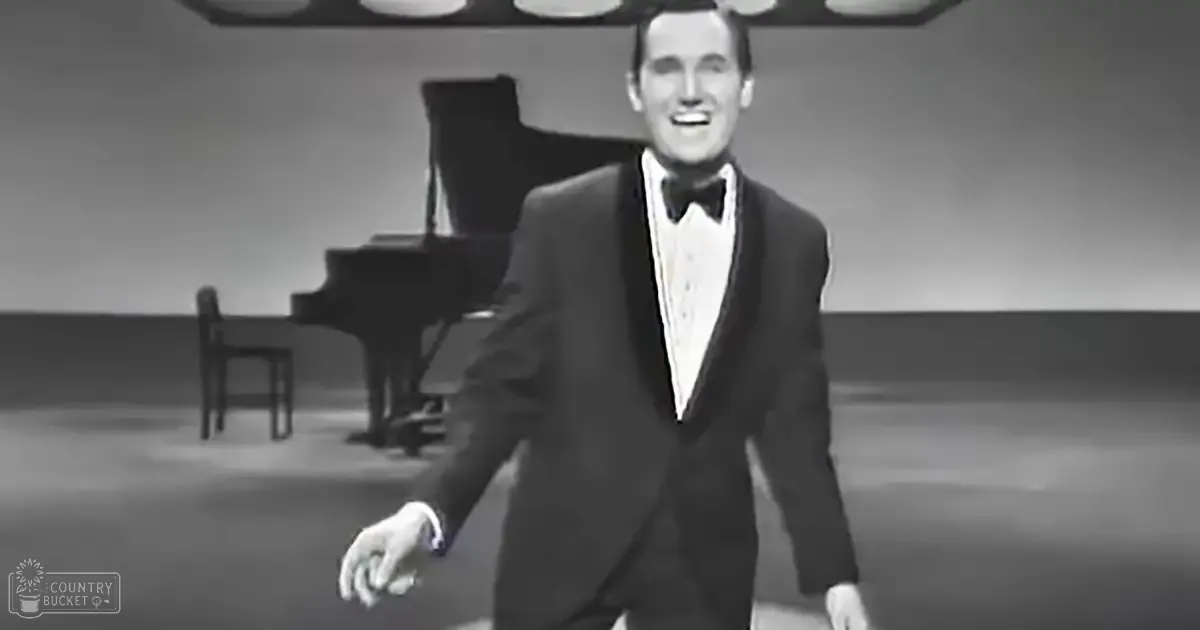 Adult male performer wearing a formal mid-20th century tuxedo and bow tie indoors on stage or in a studio with grand piano, black-and-white photograph.