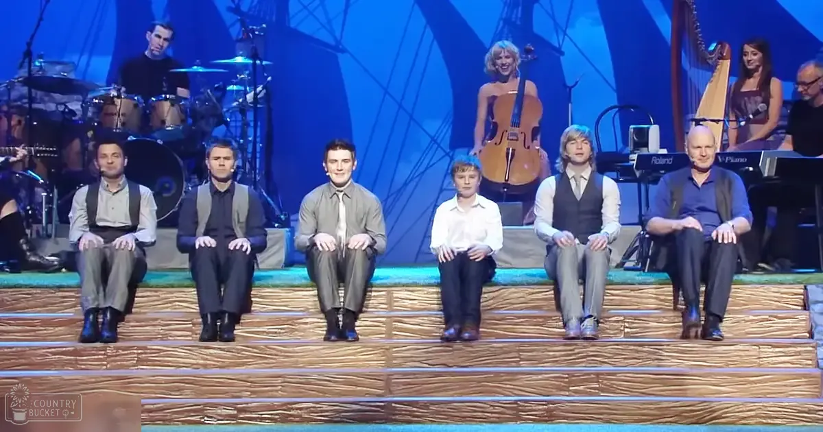 Performers on stage with a drum set, cello player, harpist, and keyboard in an indoor theater with blue backdrop and abstract geometric shapes.