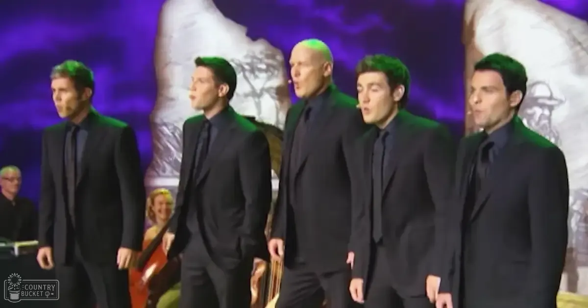Young adult male vocal group performing in an indoor concert setting with purple stage lighting, large screen displaying abstract images, orchestra seated on left side.
