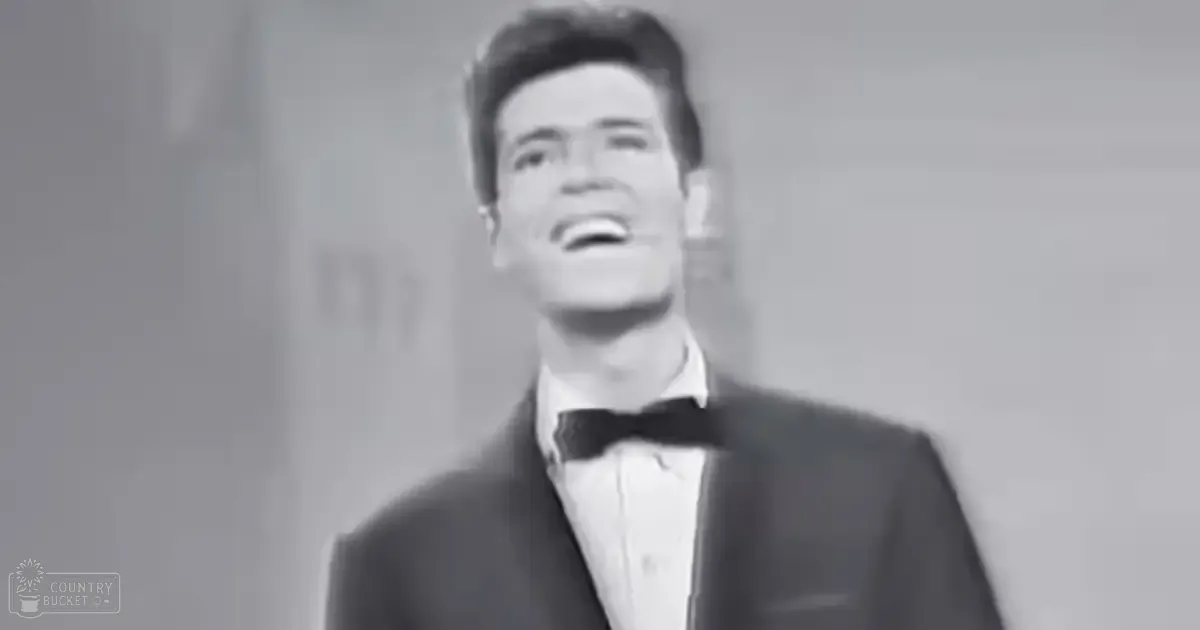 A young adult male wearing a formal suit with a bow tie, standing in an indoor setting with neutral-colored walls.