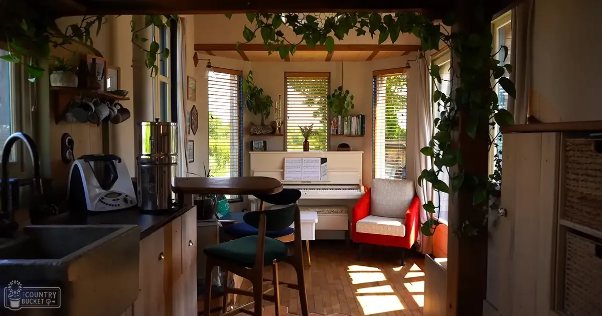 A cozy indoor space with a piano, dining table, kitchen counter, and shelves of books and decorative items. Natural light streams through large windows in spring or summer.