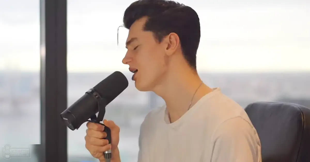 A young adult male wearing a white t-shirt and holding a microphone in an indoor setting with large windows.