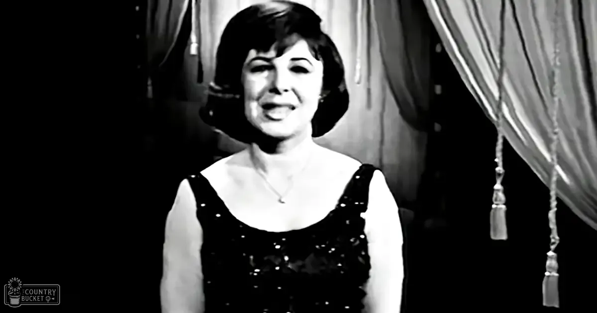 An adult female wearing a black sleeveless dress with sequined details stands indoors under dramatic stage lighting.