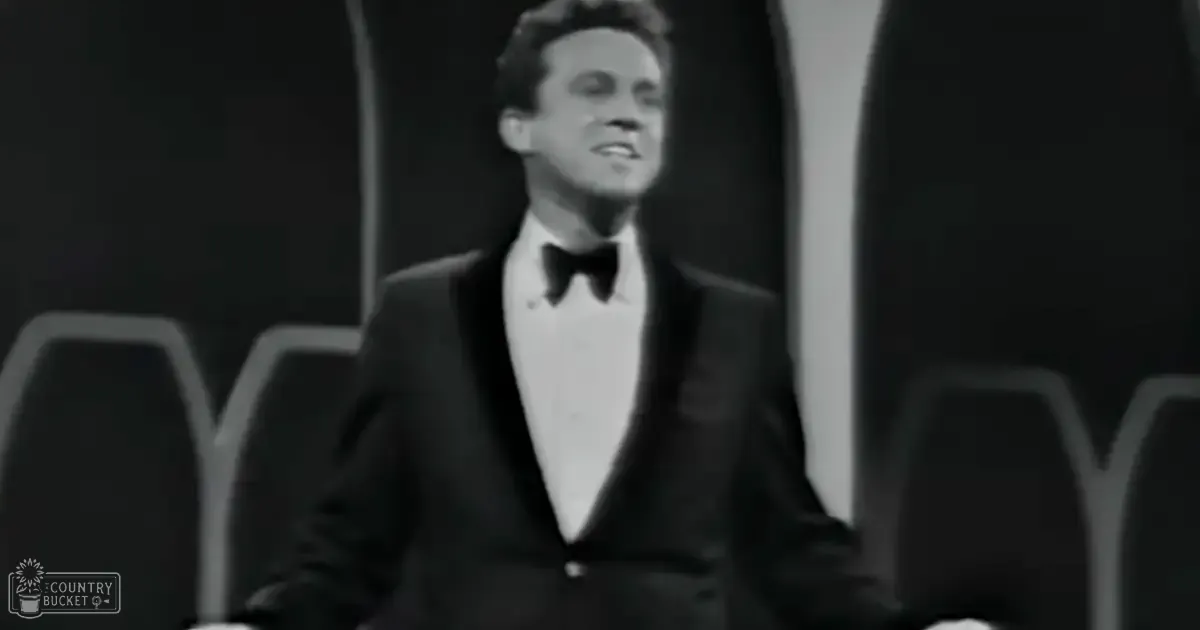 A young adult male wearing a classic fit tuxedo with a collared shirt, bow tie, and long sleeves made of silk or satin stands on an indoor stage with chairs and curtains.
