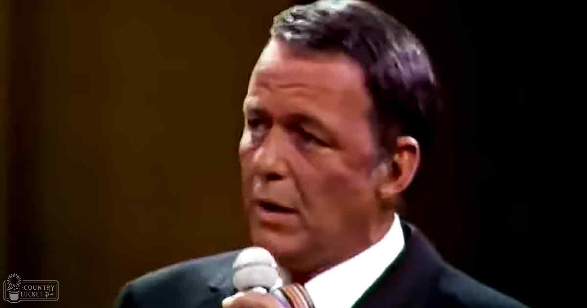 A middle-aged adult male in a formal dark suit and white shirt stands under spotlighting against a dark background with 'Madly Odd' text visible, suggesting a performance or speech context.