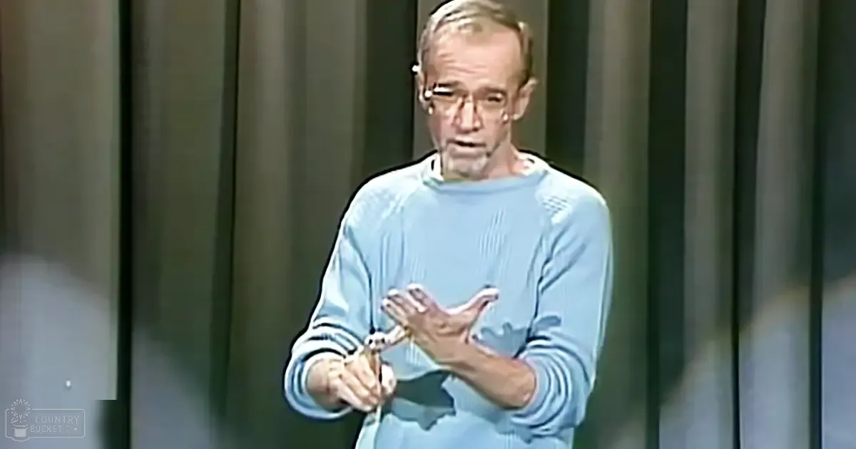 An older adult male in a mid-20th-century light blue sweater with ribbed cuffs and neckline, wearing glasses, holding a microphone against dark vertical blinds or curtains indicating an indoor performance space.
