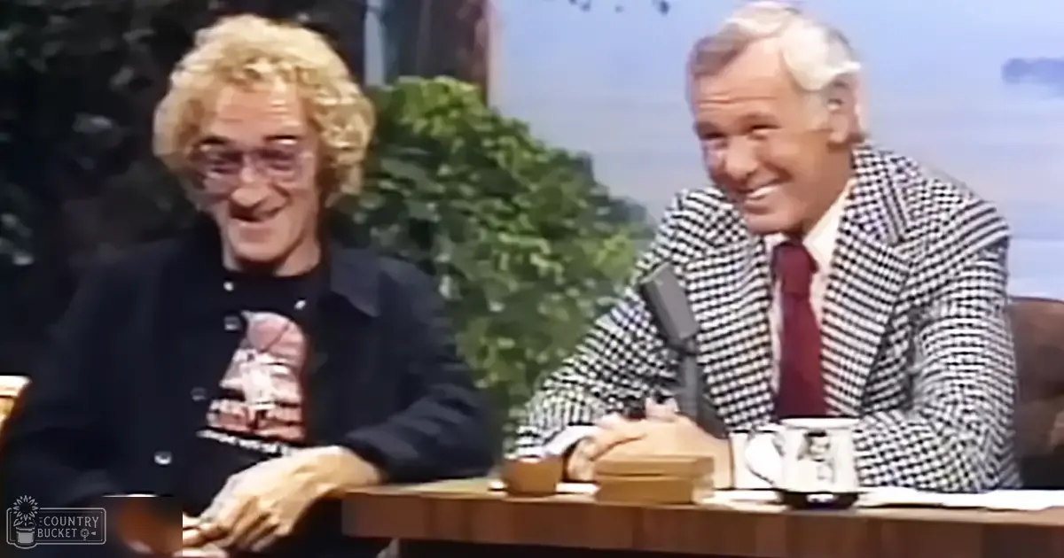 A talk show host and guest in a late-night studio setting, with one wearing a black jacket over a graphic t-shirt with an image and text; another in a checkered blazer over a white shirt with a red tie. The scene includes furniture like a wooden table with a mug displaying a person's face printed on it.