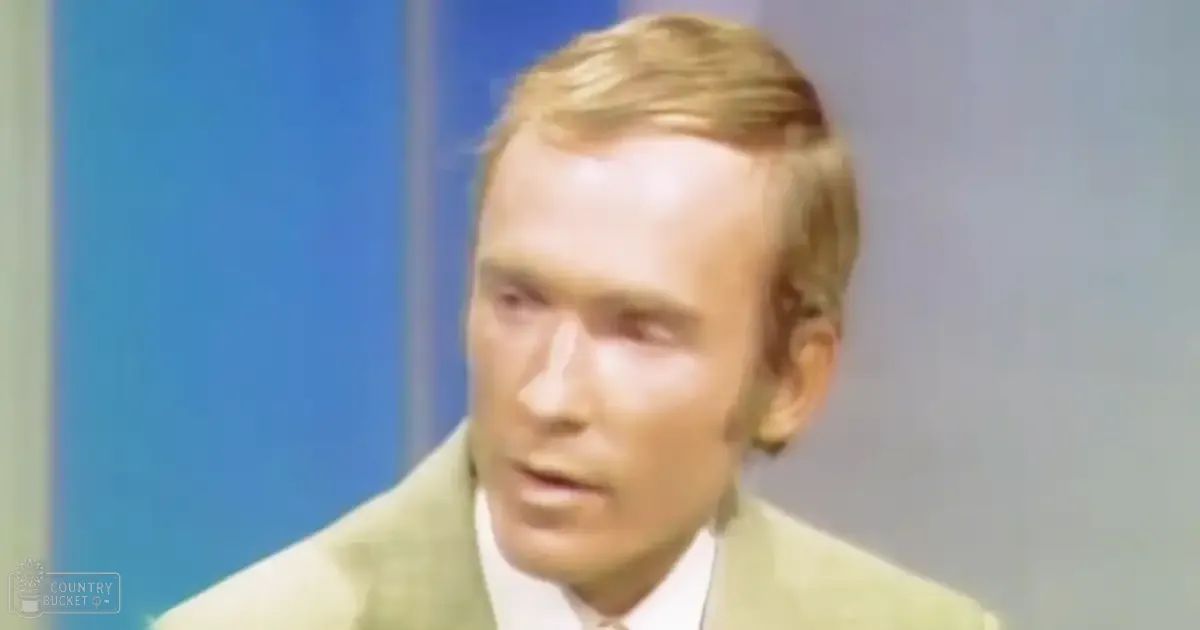 A middle-aged adult male wearing a light green suit jacket over a white collared shirt speaks against a blue wall backdrop, suggesting an indoor studio or office environment.