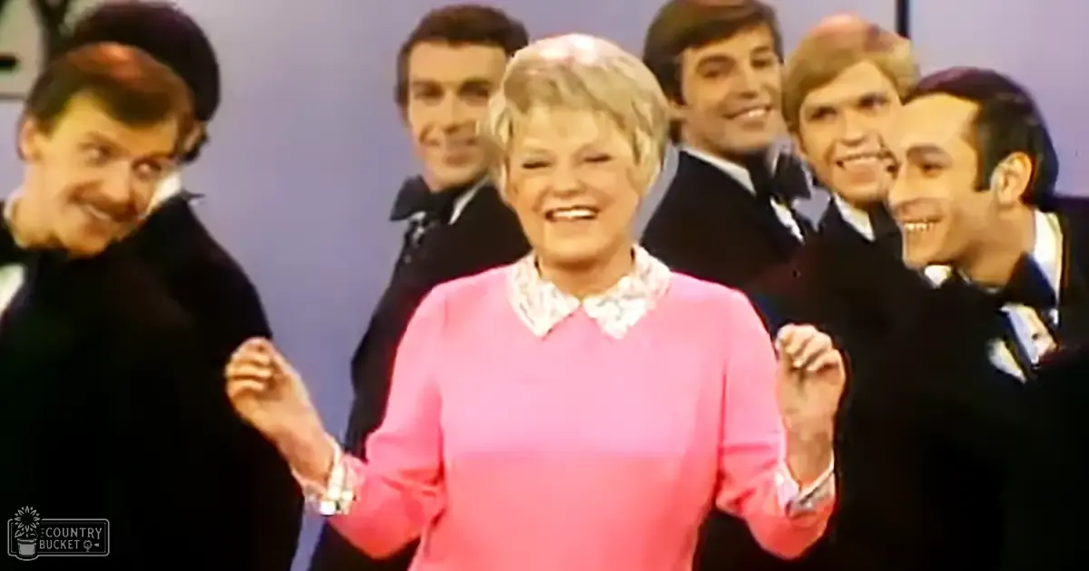 A middle-aged adult female wearing a pink sweater with a white lace collar stands at the forefront of an indoor event or performance setting.