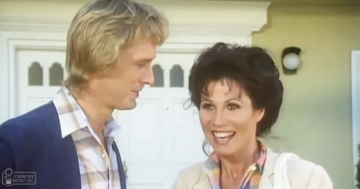 A middle-aged woman wearing a light-colored blazer interacts with a young adult man in front of a residential house during late afternoon daylight.