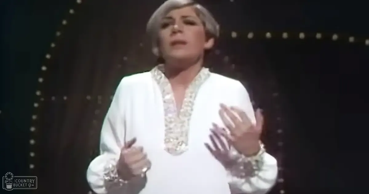 An adult female performer in an indoor setting, possibly a stage or studio, wearing a white silk blouse with intricate embroidery and cuffs. The background suggests a theatrical environment with dark tones and decorative elements.