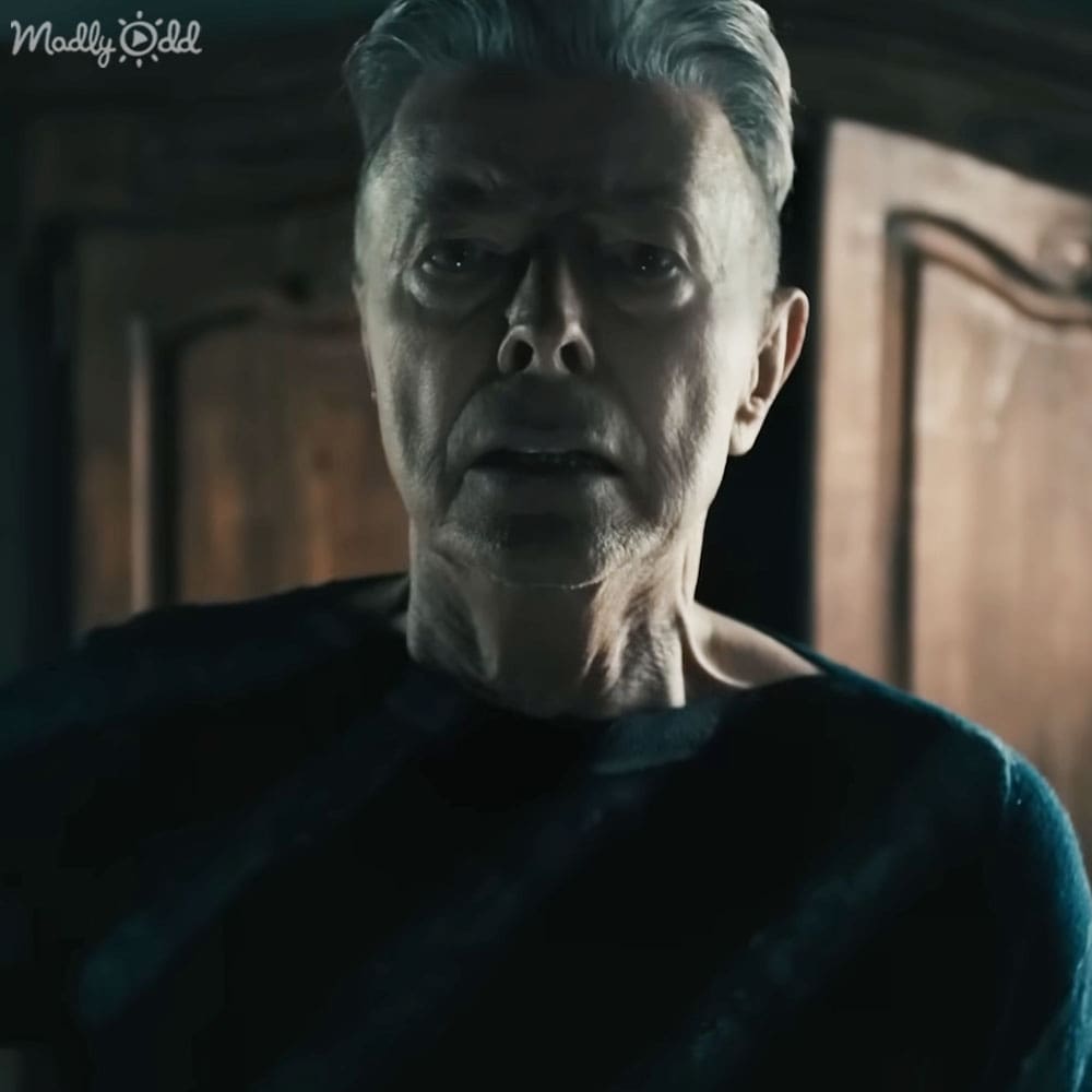 An older adult male in a dimly lit room with ornate wooden paneling, wearing a formal or historical dark-colored garment.