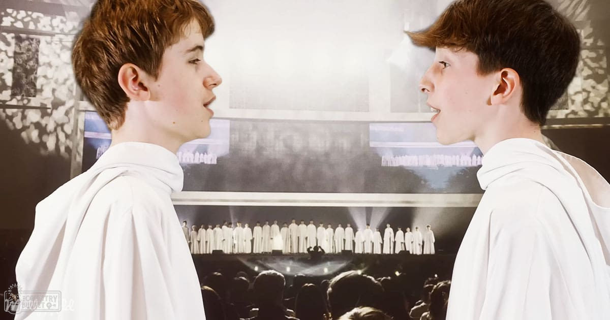 Two young adults in white choir robes performing indoors in a concert hall setting with bright stage lighting and an audience.