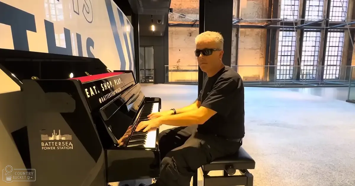 An adult male wearing a black T-shirt and sunglasses plays a piano at Battersea Power Station, with industrial scaffolding and construction in the background.