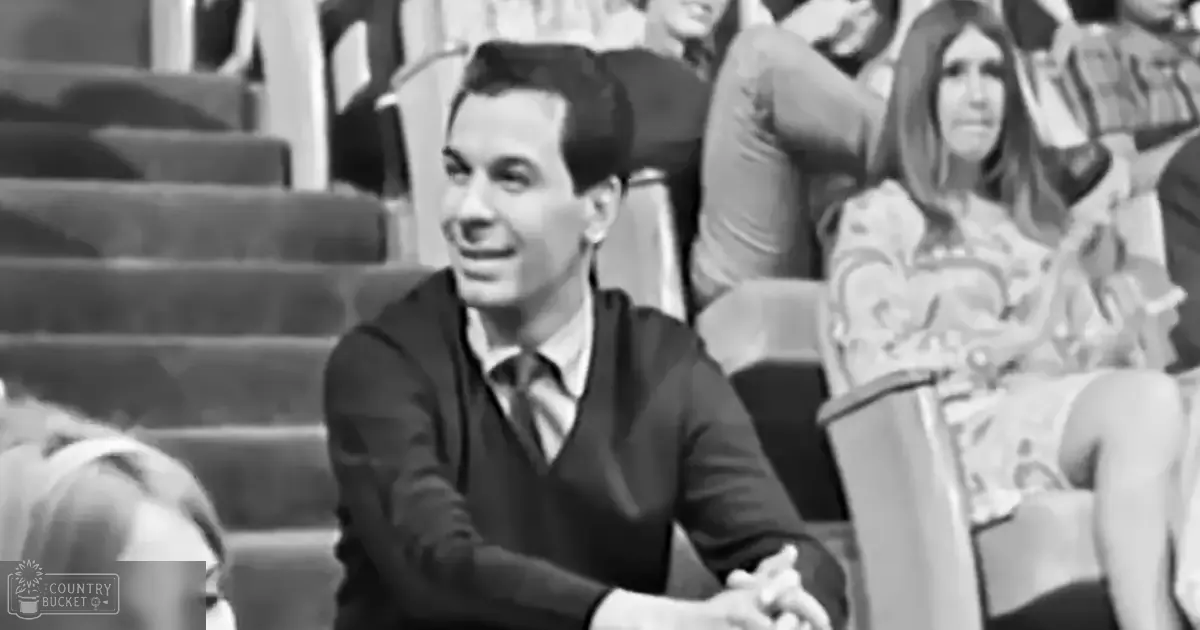 A young adult male in a modern, possibly late 20th-century style shirt and sweater vest stands in an auditorium setting with tiered seating, while an adult female in a patterned dress and child in light-colored clothing are also present.
