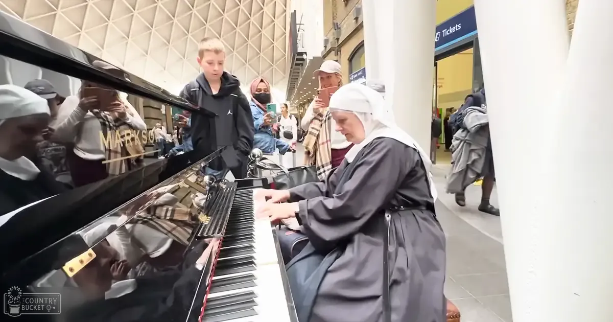A nun playing a piano in an indoor public space with modern architectural design; background includes a child near the piano, several middle-aged women wearing various types of clothing and accessories.