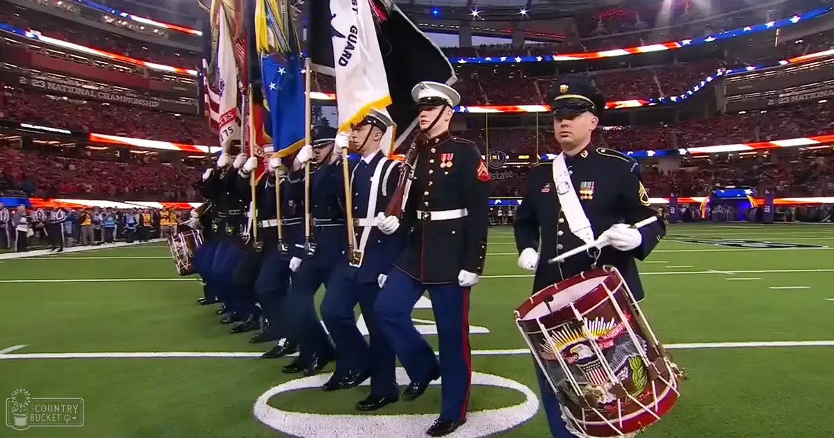 Military parade at a large stadium during an event, with adult males in dark blue military uniforms with white gloves, white hats, and black shoes; flags and drums are notable objects; large indoor sports arena setting with artificial lighting and rows of spectators.
