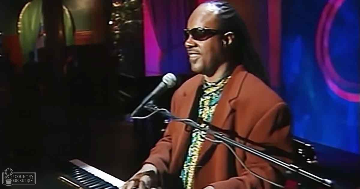 An adult male musician performing, wearing a brown blazer over a patterned shirt with a high collar and long sleeves. Setting includes microphone stand, microphones near his mouth, piano keyboard, dark blue and purple curtains or stage decorations, 'Madly Odd' logo.