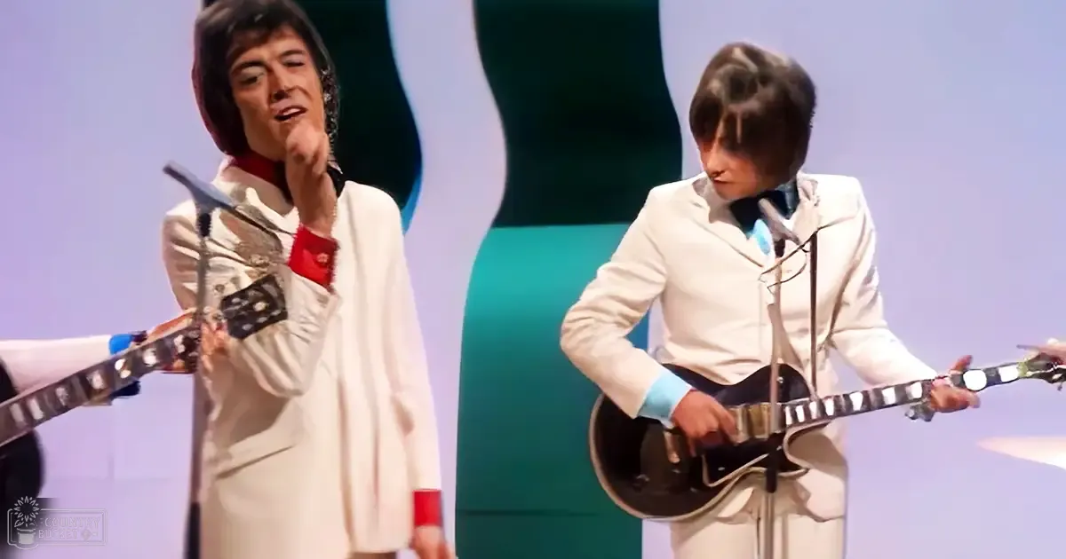 A stage setting with two young adult male performers wearing matching white suits, standing side by side against a simple green and white backdrop under bright artificial lighting.