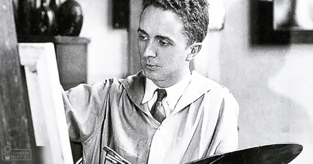 A young adult male artist wearing a loose-fitting white smock over a dark shirt, working in an art studio with various objects on shelves and framed artwork hanging behind him.