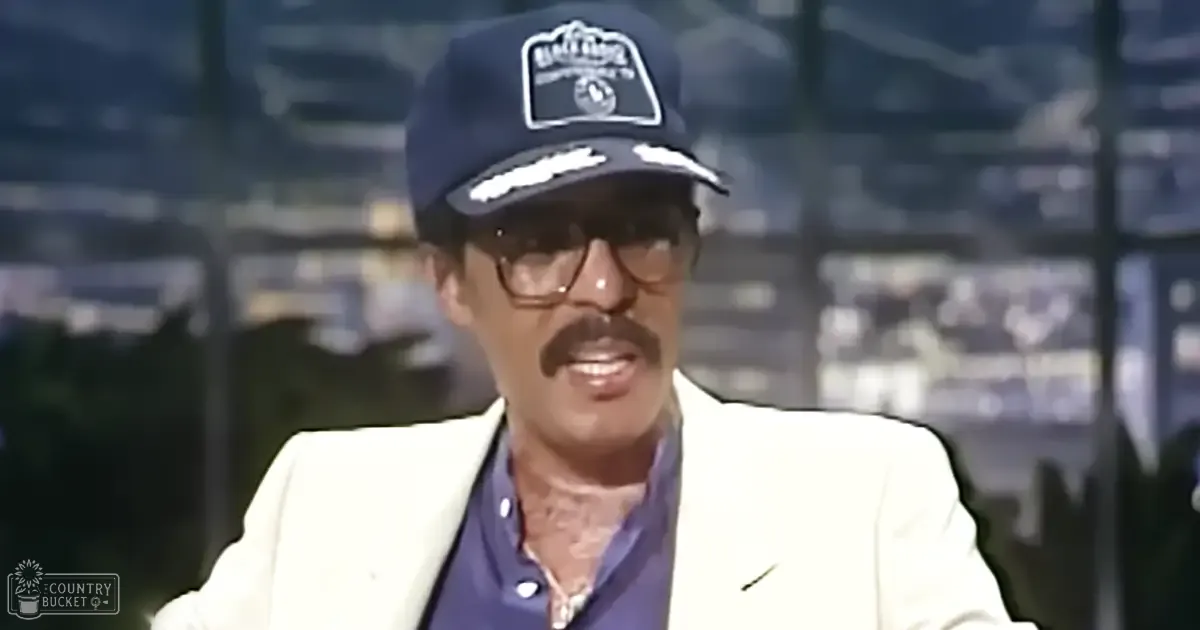 An older adult male wearing a white blazer, dark blue collared shirt, navy baseball cap labeled 'Baker's', and glasses in an indoor sports arena setting.