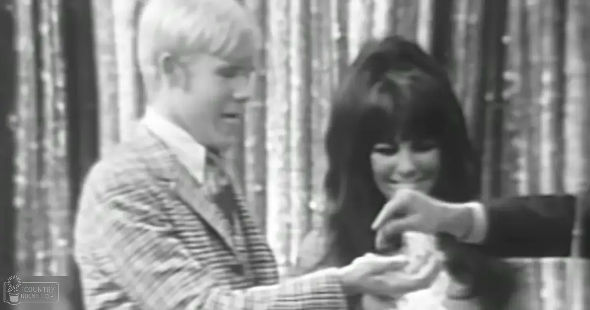 A young adult couple indoors; male wears a striped blazer over collared shirt and tie from the mid-20th century; female in dark V-neck dress typical of late 1960s or early 1970s fashion style, with vertical curtains suggesting formal setting like restaurant or event venue