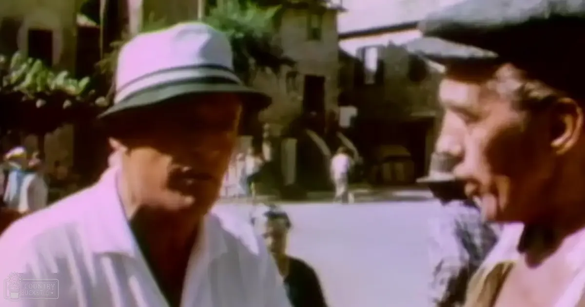 Two older adult males wearing button-up shirts and fedoras stand outdoors on a street with mid-20th-century architectural elements.