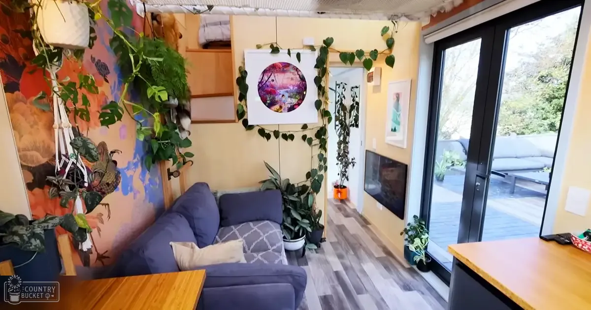 Cozy living room scene with blue sofa, wooden coffee table, wall-mounted TV, hanging plants, wall art, sliding glass door leading to an outdoor patio area in a residential space during daytime.