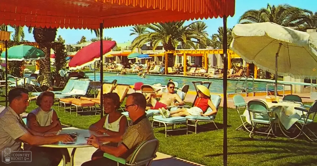 Adults enjoying leisure time at a resort-style outdoor poolside with lounge chairs, palm trees, and colorful umbrellas in the background.