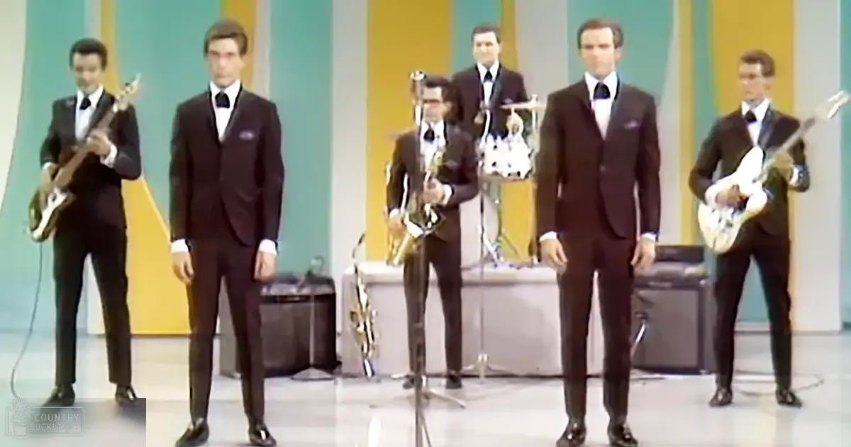 Musical performance scene indoors with vertical stripes in yellow, teal, and white backdrop; musical instruments like guitars and saxophone visible on stage.